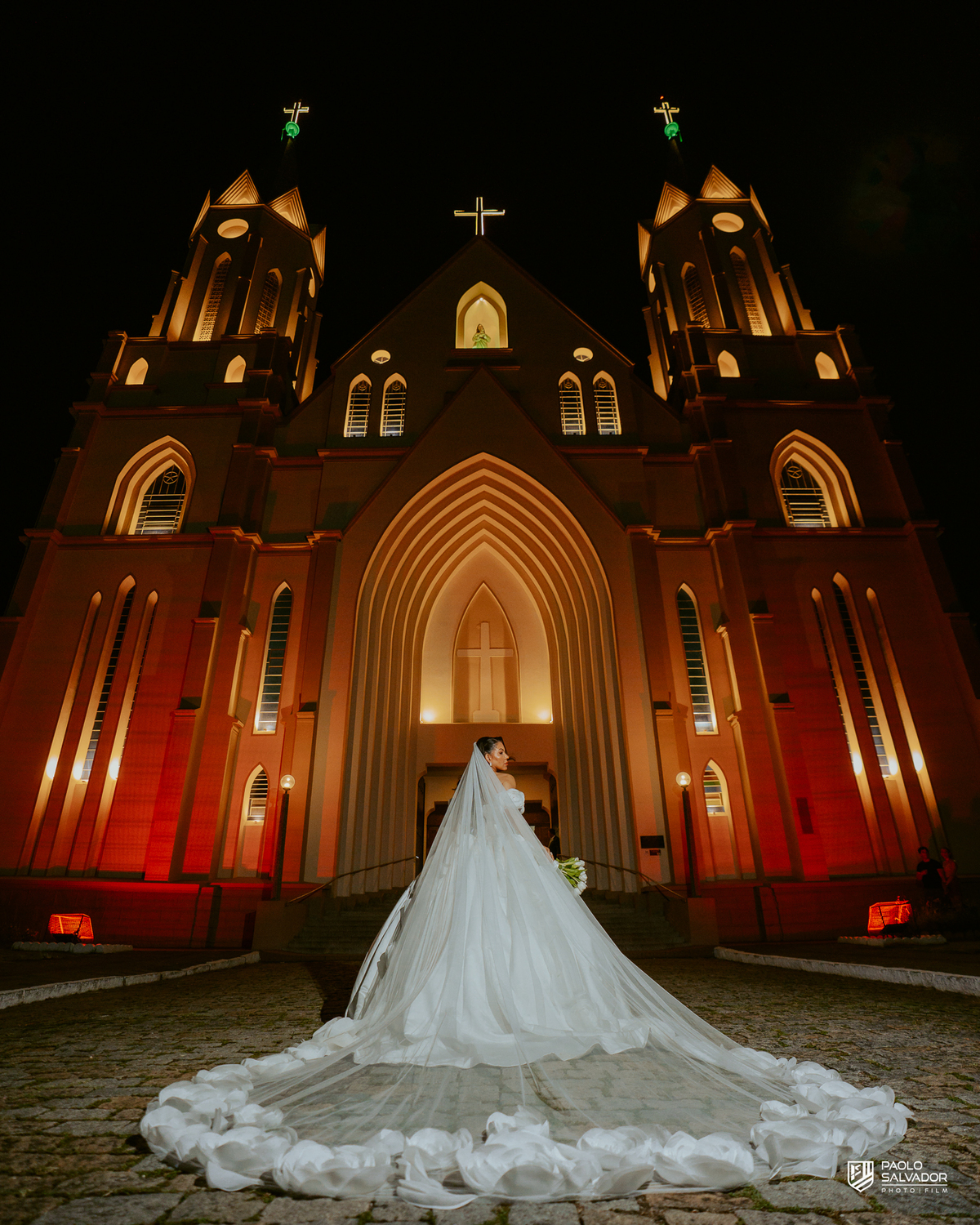 Noiva com véu sendo ajustado pela equipe em frente à Igreja Santa Inês em Indaial à noite, preparação para ensaio pós-cerimônia, fotografia de casamento noturno