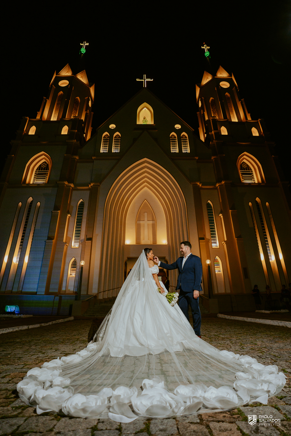 Noiva com véu sendo ajustado pela equipe em frente à Igreja Santa Inês em Indaial à noite, preparação para ensaio pós-cerimônia, fotografia de casamento noturno