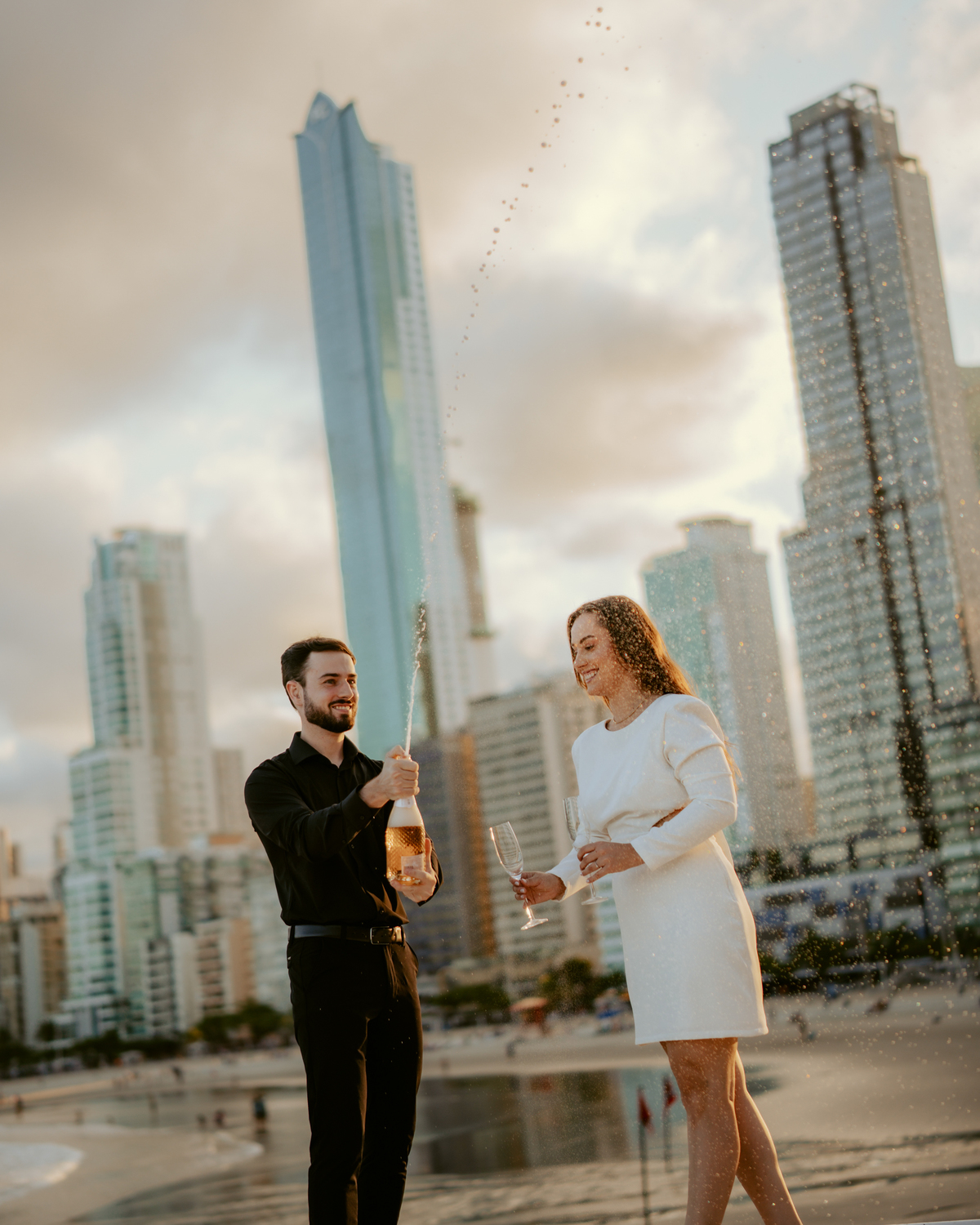 Gabi e Henrique em ensaio pré-wedding na FG Big Wheel de Balneário Camboriú, casal caminhando na orla com vista para o mar e cidade, estilo urbano contemporâneo com pôr do sol e composição cinematográfica