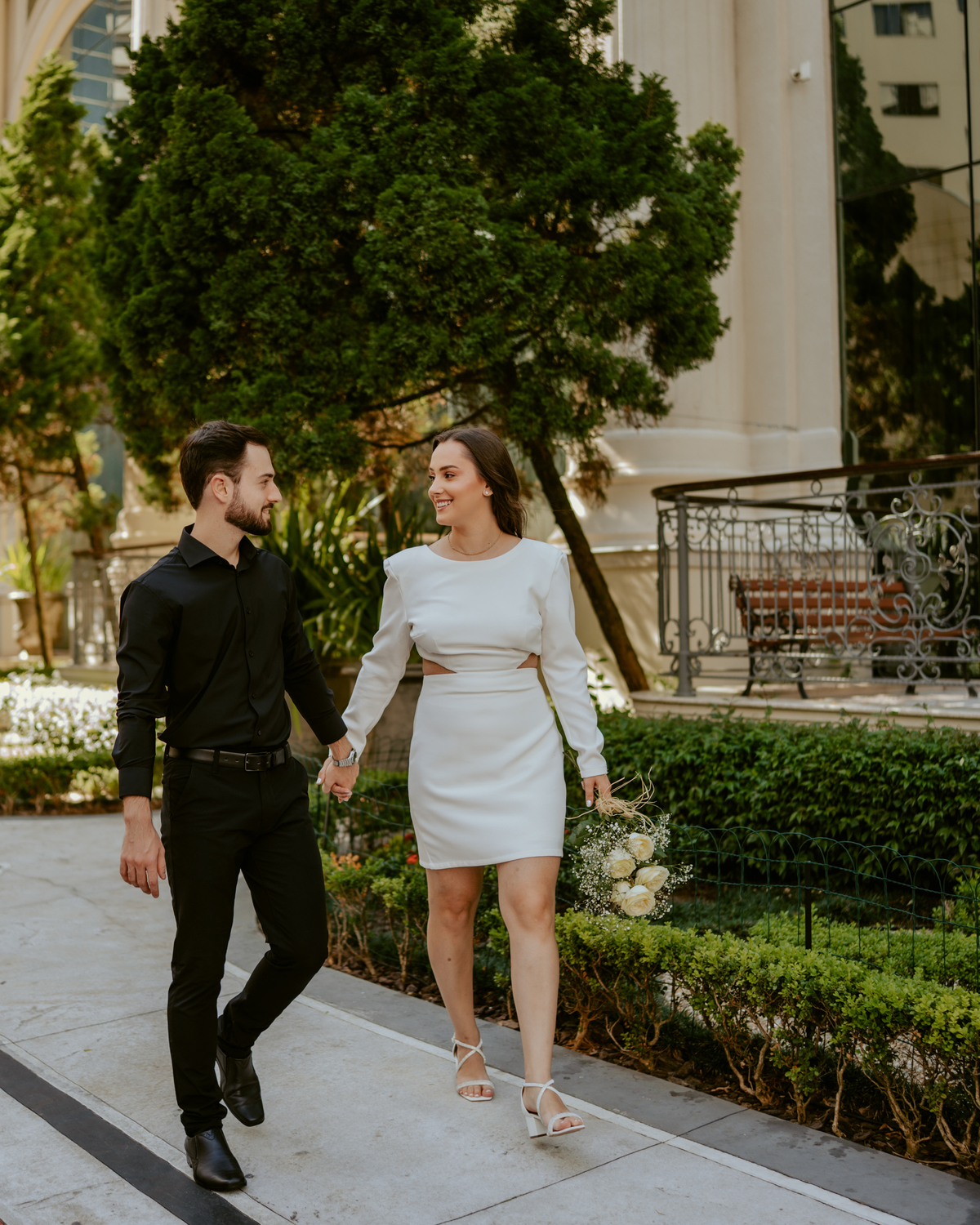Ensaio pré-wedding moderno em Balneário Camboriú com casal elegante caminhando de mãos dadas em frente a prédio com arquitetura clássica contemporânea, look preto e branco sofisticado, fotografia urbana romântica ao ar livre
