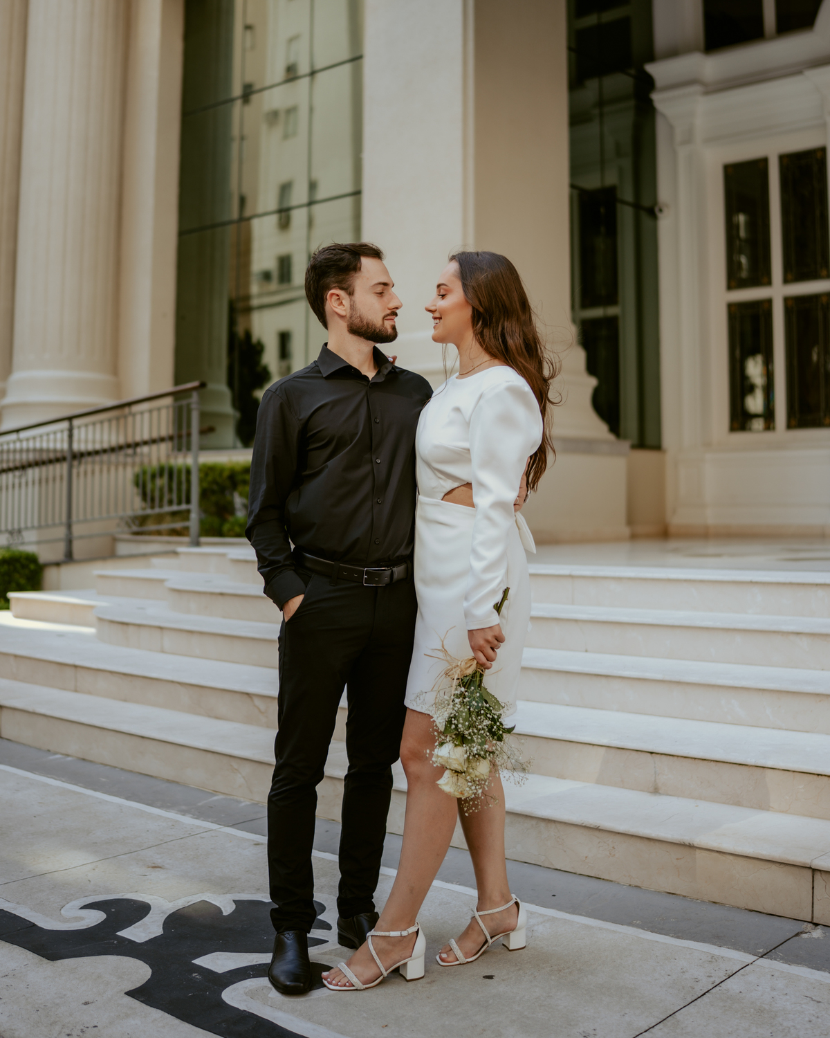 Ensaio pré-wedding moderno em Balneário Camboriú com casal elegante caminhando de mãos dadas em frente a prédio com arquitetura clássica contemporânea, look preto e branco sofisticado, fotografia urbana romântica ao ar livre