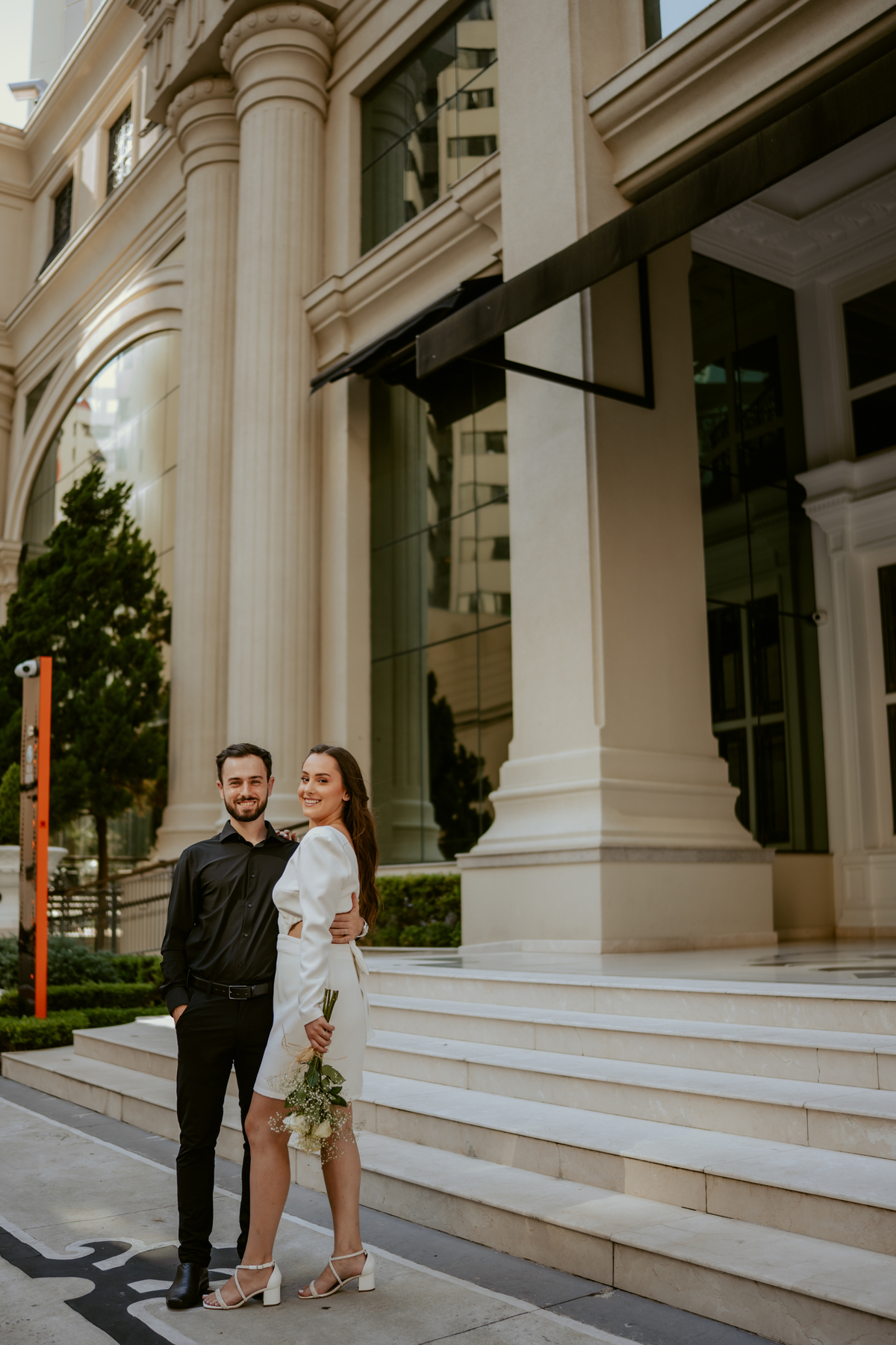 Ensaio pré-wedding moderno em Balneário Camboriú com casal elegante caminhando de mãos dadas em frente a prédio com arquitetura clássica contemporânea, look preto e branco sofisticado, fotografia urbana romântica ao ar livre