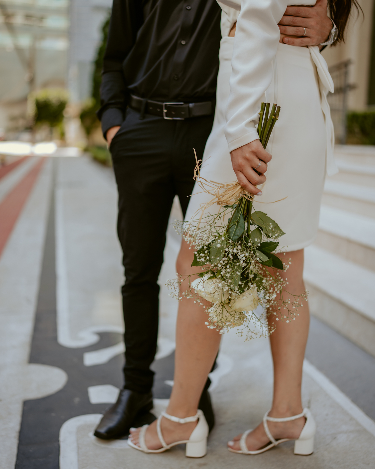 Ensaio pré-wedding moderno em Balneário Camboriú com casal elegante caminhando de mãos dadas em frente a prédio com arquitetura clássica contemporânea, look preto e branco sofisticado, fotografia urbana romântica ao ar livre