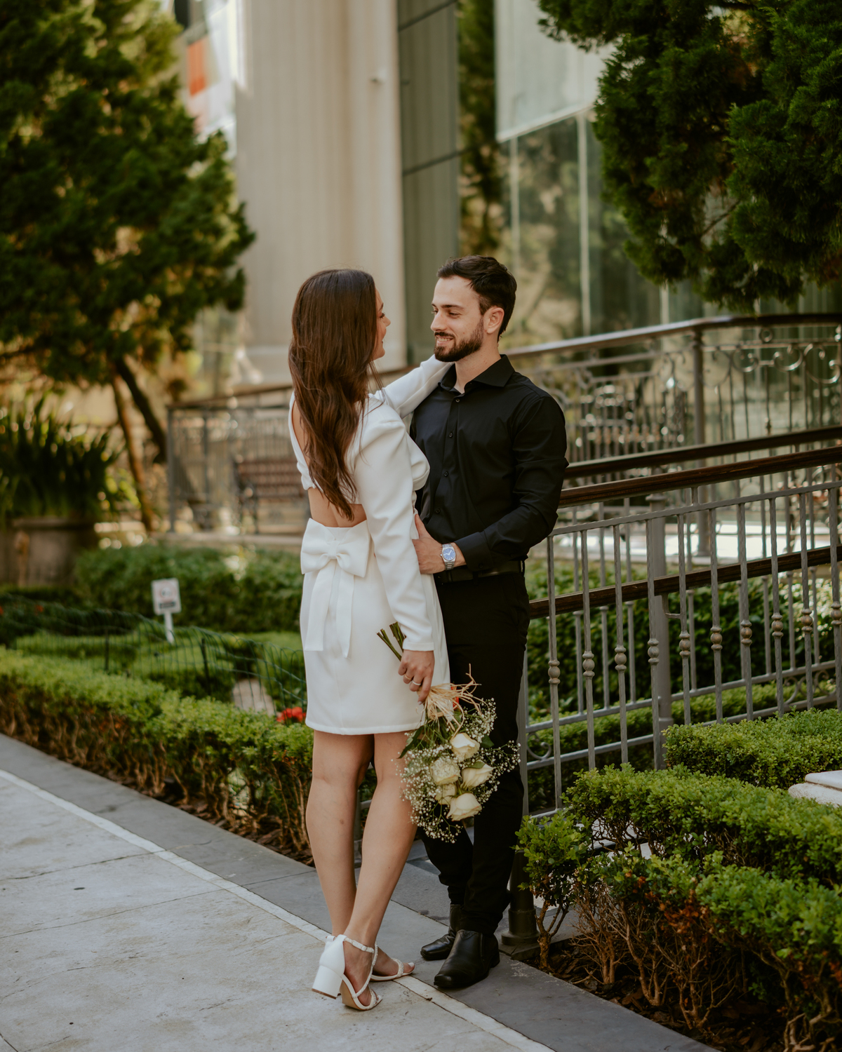Ensaio pré-wedding moderno em Balneário Camboriú com casal elegante caminhando de mãos dadas em frente a prédio com arquitetura clássica contemporânea, look preto e branco sofisticado, fotografia urbana romântica ao ar livre