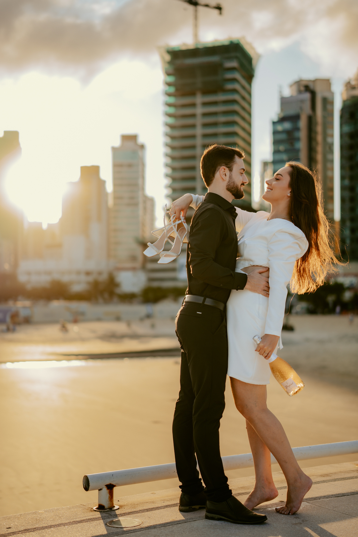 Ensaio pré-wedding na orla de Balneário Camboriú próximo à roda gigante FG Big Wheel, casal sorrindo e brindando com champagne no pier, fotografia espontânea, moderna e romântica com skyline e luz dourada