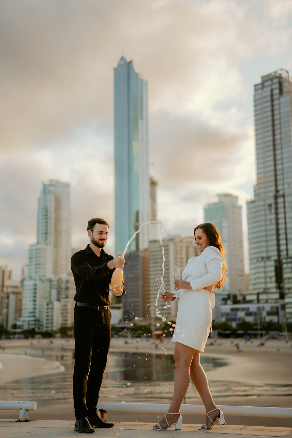 Gabi e Henrique em ensaio pré-wedding na FG Big Wheel de Balneário Camboriú, casal caminhando na orla com vista para o mar e cidade, estilo urbano contemporâneo com pôr do sol e composição cinematográfica