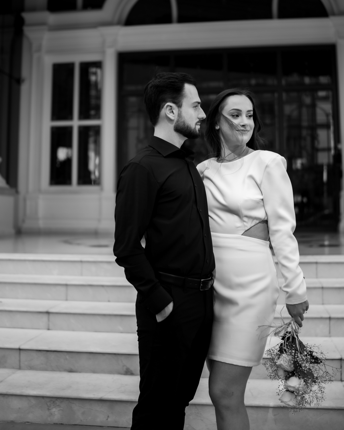 Ensaio pré-wedding moderno em Balneário Camboriú com casal elegante caminhando de mãos dadas em frente a prédio com arquitetura clássica contemporânea, look preto e branco sofisticado, fotografia urbana romântica ao ar livre