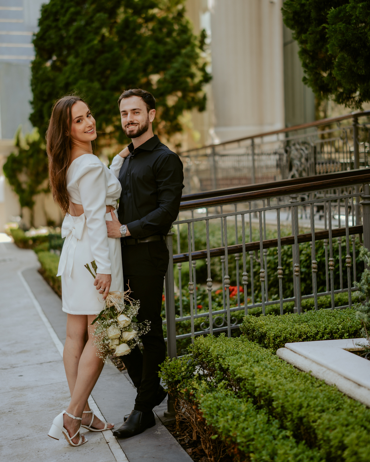 Ensaio pré-wedding moderno em Balneário Camboriú com casal elegante caminhando de mãos dadas em frente a prédio com arquitetura clássica contemporânea, look preto e branco sofisticado, fotografia urbana romântica ao ar livre