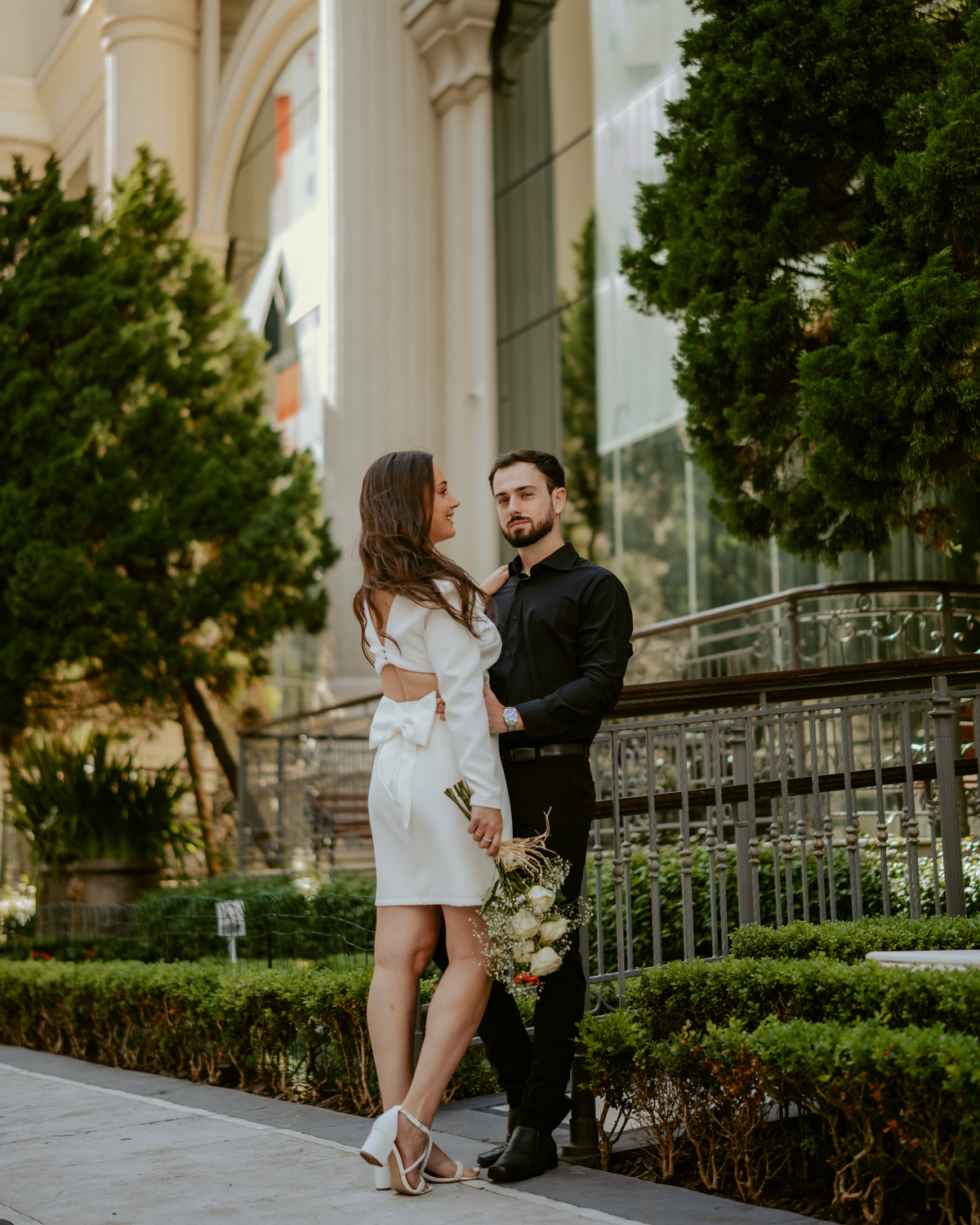 Ensaio pré-wedding moderno em Balneário Camboriú com casal elegante caminhando de mãos dadas em frente a prédio com arquitetura clássica contemporânea, look preto e branco sofisticado, fotografia urbana romântica ao ar livre