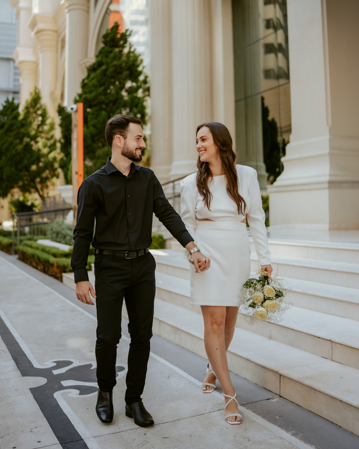 Ensaio pré-wedding moderno em Balneário Camboriú com casal elegante caminhando de mãos dadas em frente a prédio com arquitetura clássica contemporânea, look preto e branco sofisticado, fotografia urbana romântica ao ar livre