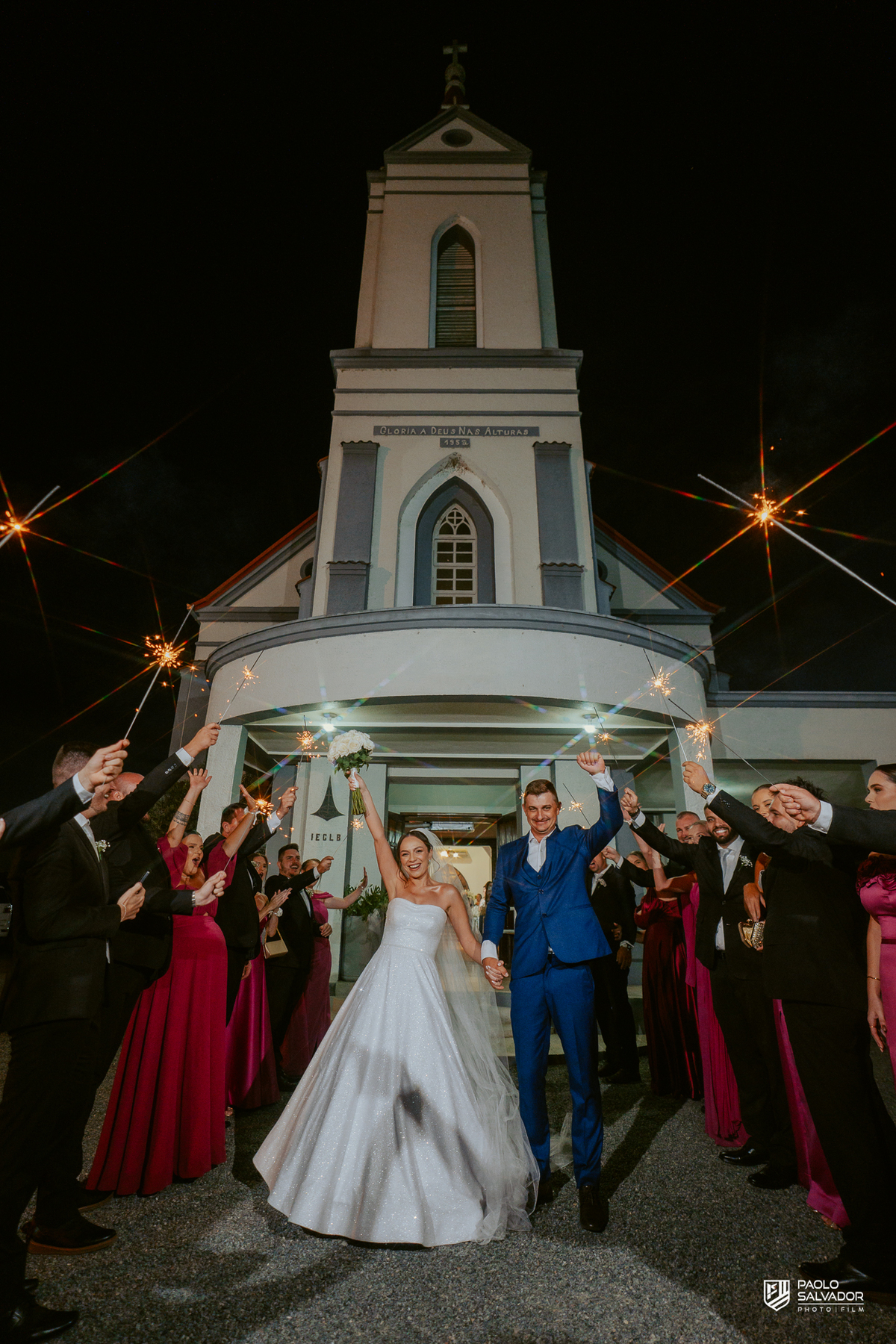 Saída dos noivos com sparkles em frente à igreja em Massaranduba, casamento Jaque e Thayro à noite, fotografia de casamento com luz criativa e momento de celebração com convidados