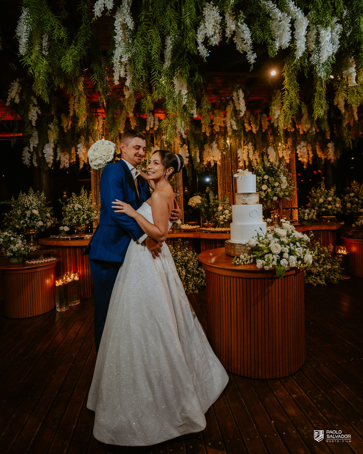 Noivos Jaque e Thayro na festa no Refúgio dos Lagos em Luiz Alves, decoração Vila das Flores, bolo e pista de dança, fotografia de casamento sofisticada em Massaranduba e região, retrato casal na mesa do bolo