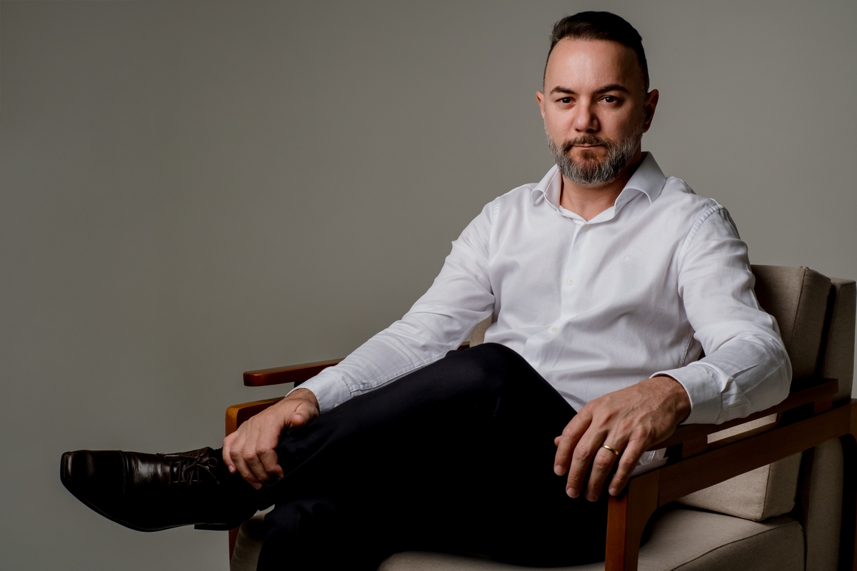 Homem de negócios sentado em poltrona de madeira com postura confiante, vestindo camisa branca e calça escura contra fundo claro.