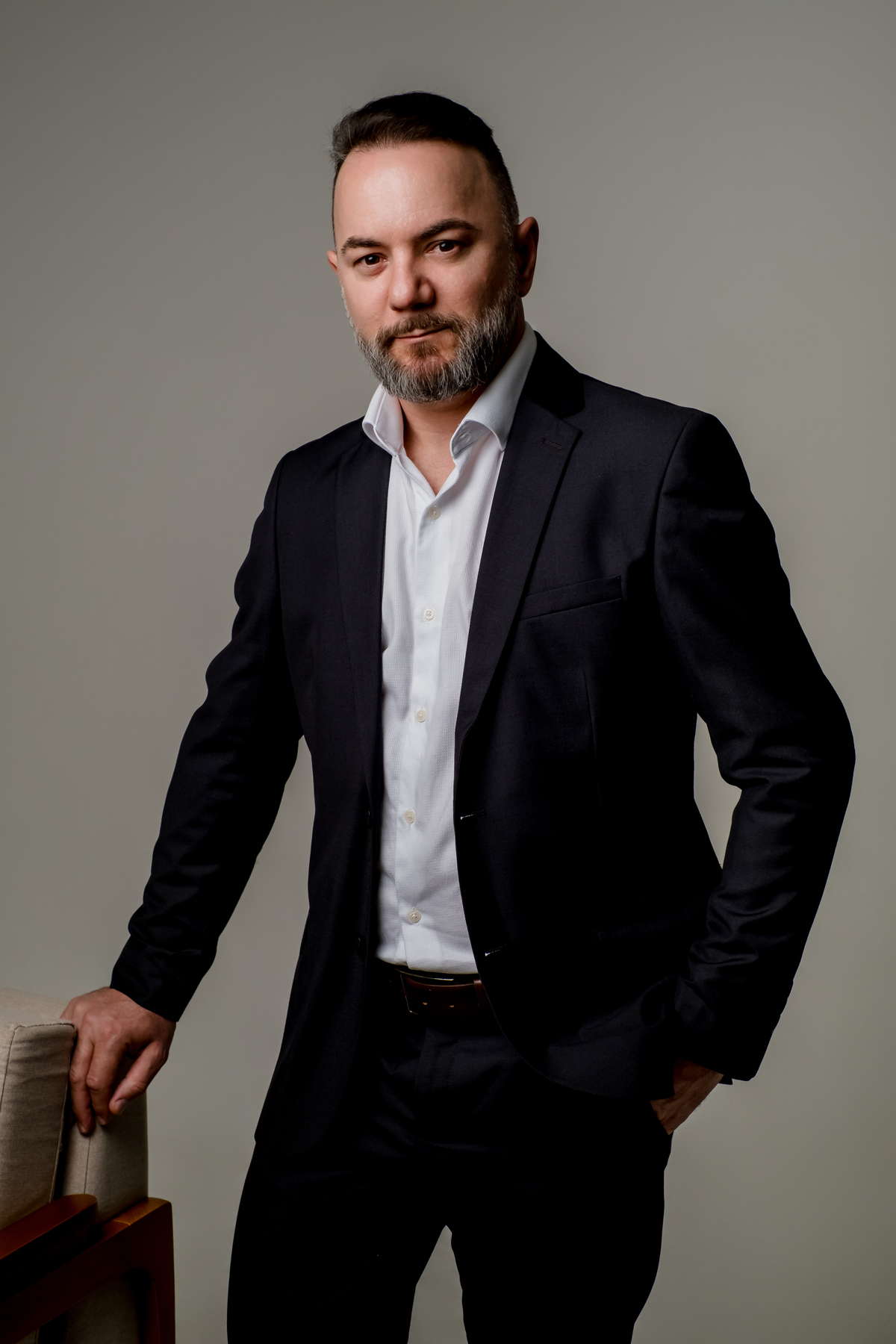 Retrato executivo de homem em pé de terno escuro, apoiado em poltrona, com olhar direto e fundo claro minimalista.