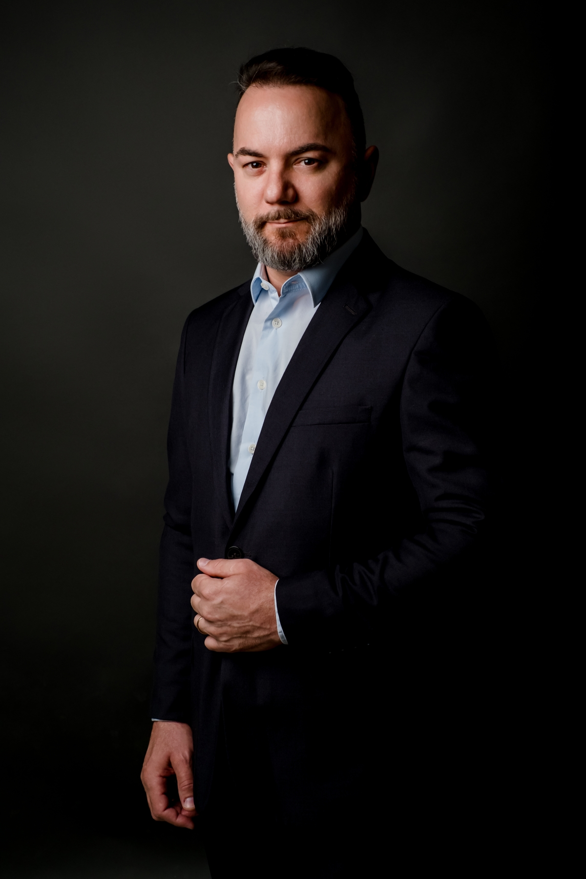 Homem em postura executiva ajustando o terno escuro, camisa azul, transmitindo elegância e prontidão contra fundo preto.