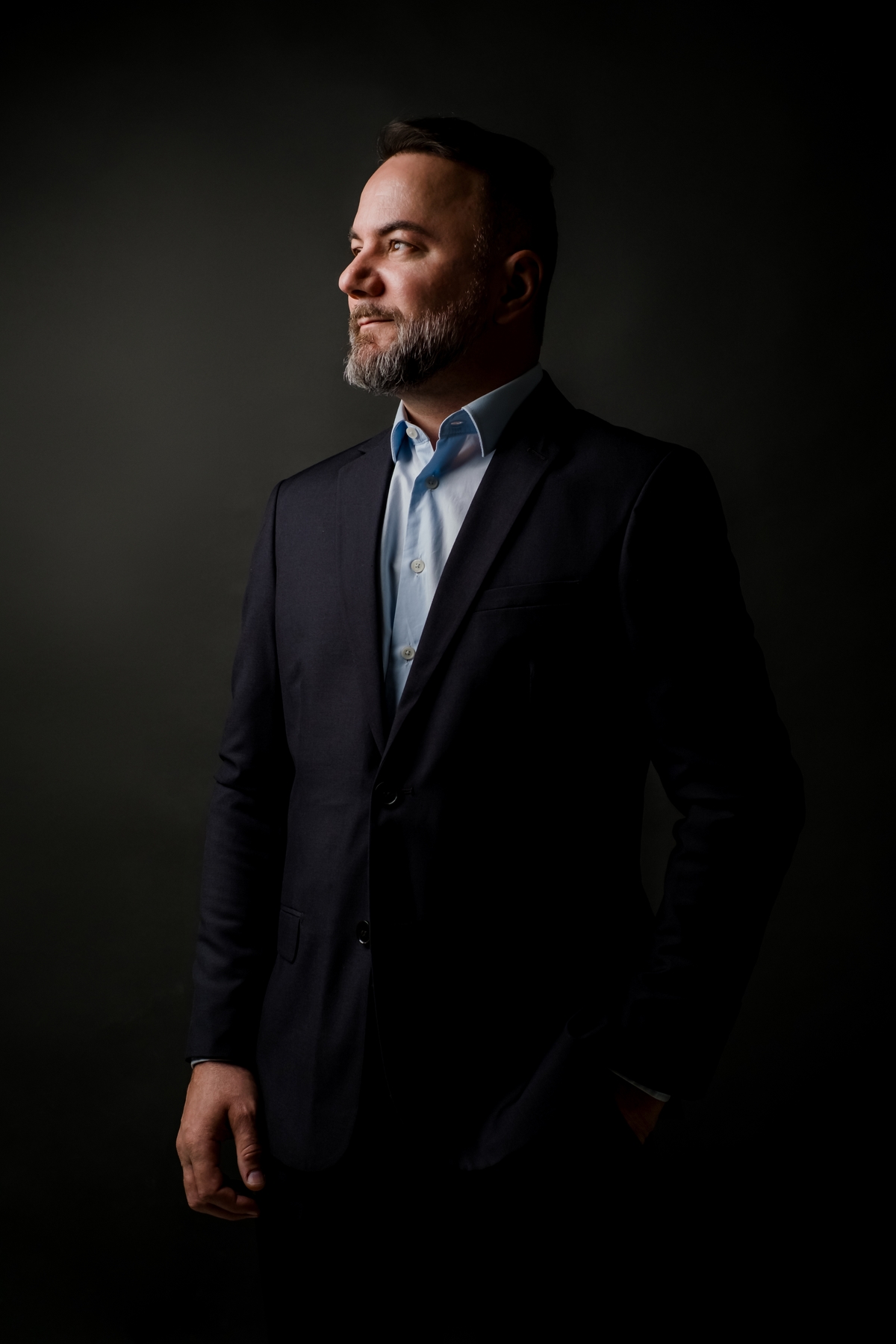 Fotografia executiva de homem de terno escuro em pé com as mãos no bolso, olhando para o lado em perfil com iluminação marcada.