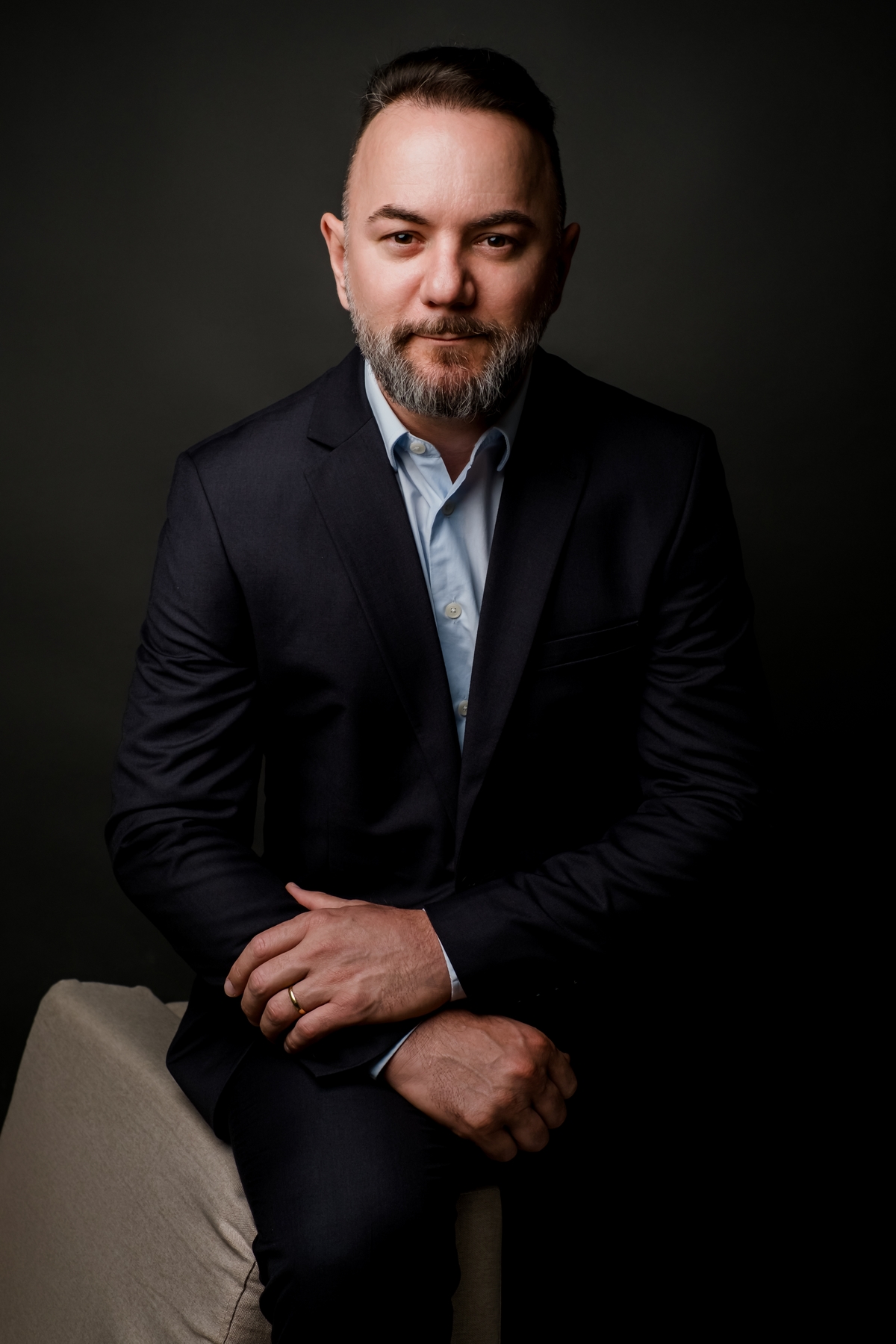 Retrato corporativo de homem sentado com as mãos apoiadas, vestindo terno escuro e olhando diretamente para a câmera, fundo preto.