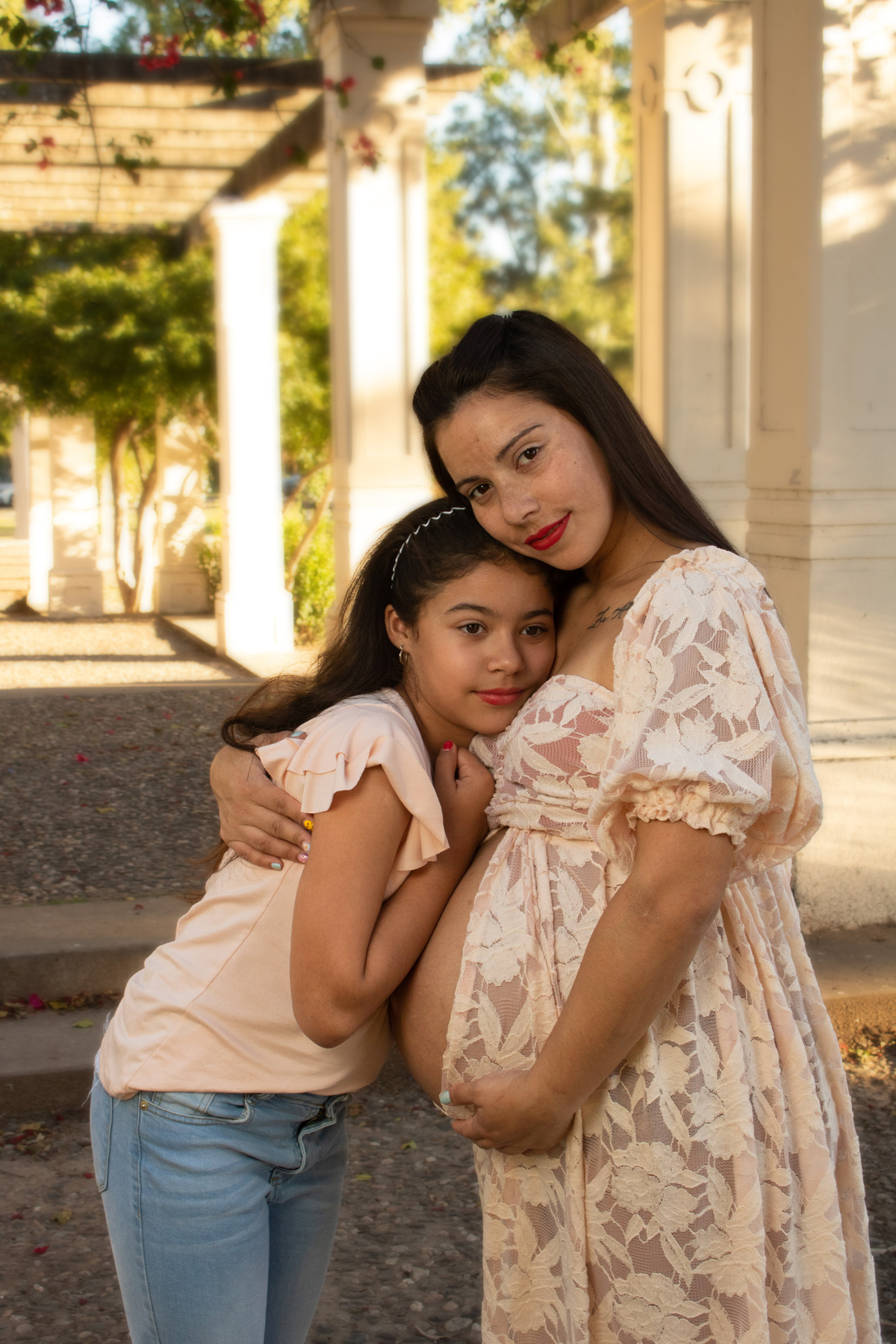 embarazada de perfil, mirando al frente, abrazada a su hija,  en una glorieta, vestido romántico