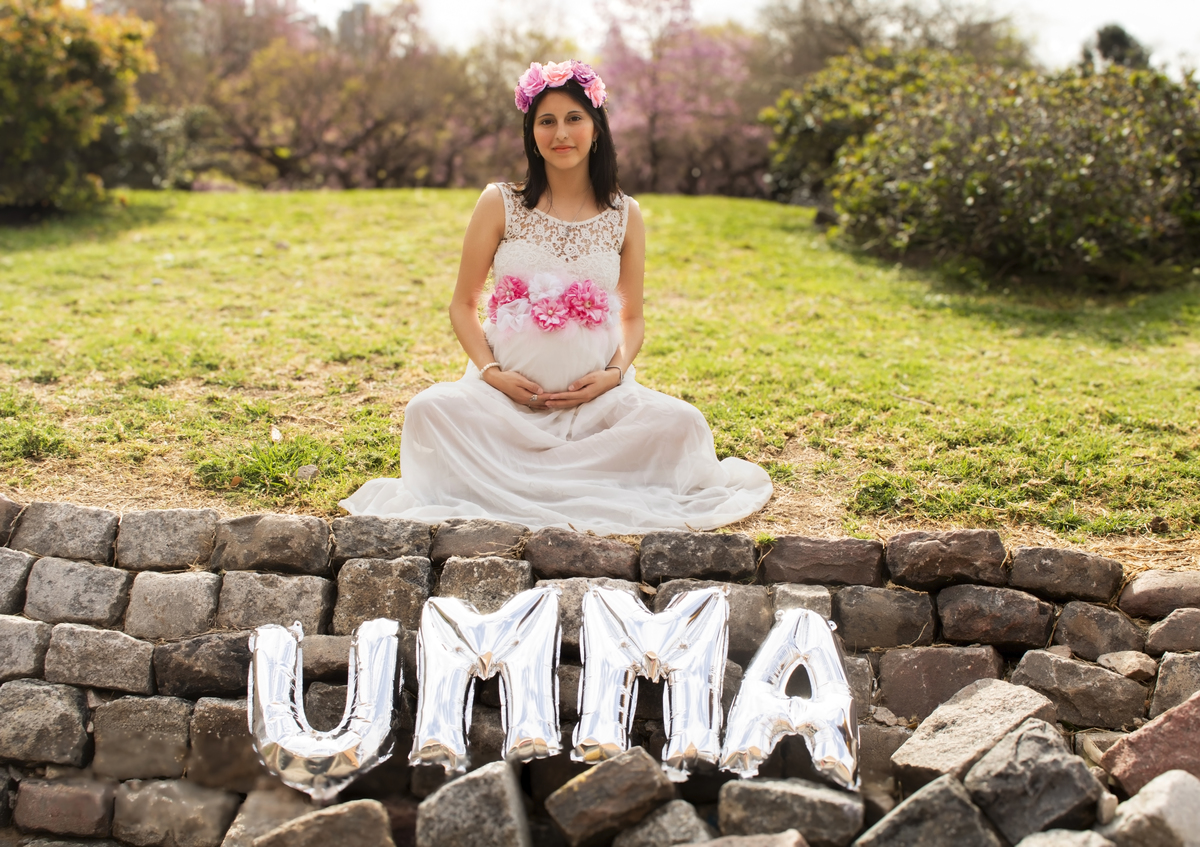 embarazada sentada sobre el césped, con globos de letras formando el nombre de su bebe, plano general