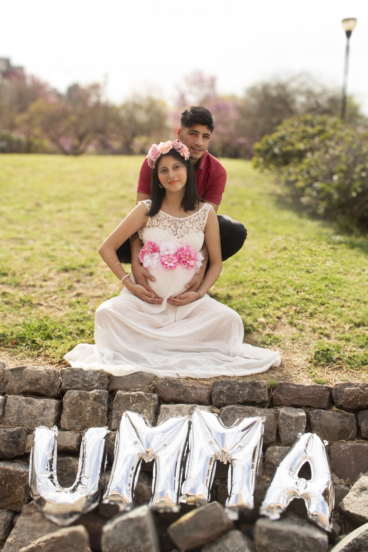 embarazada sentada sobre el césped, con su pareja,  con globos de letras formando el nombre de su bebe, 