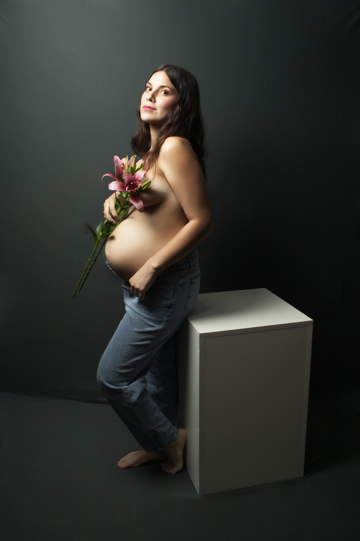 embarazada con el torso desnudo cubriéndose con flores de lilium apoyada en un cubo blanco