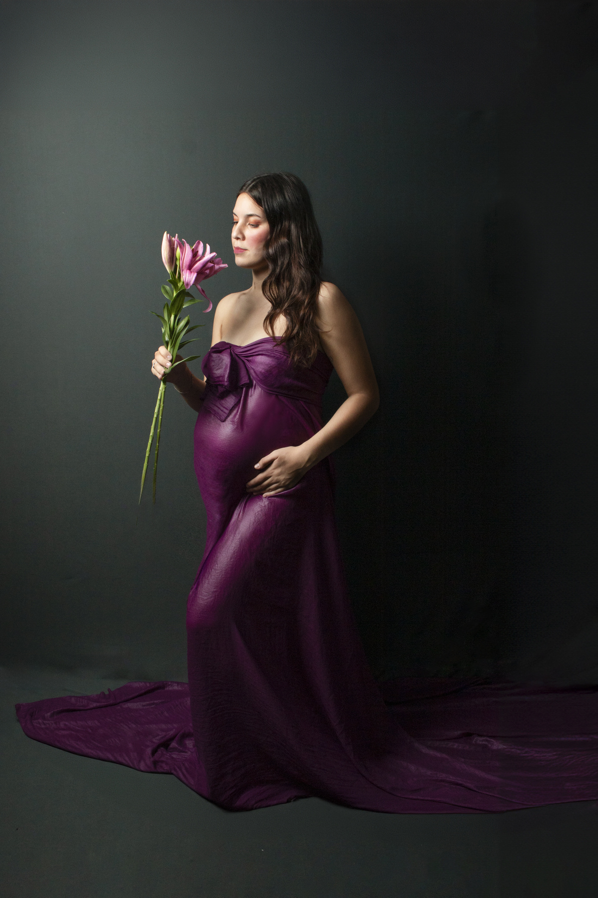 Embarazada con vestido largo con cola color violeta y flores de lilium, con fondo oscuro