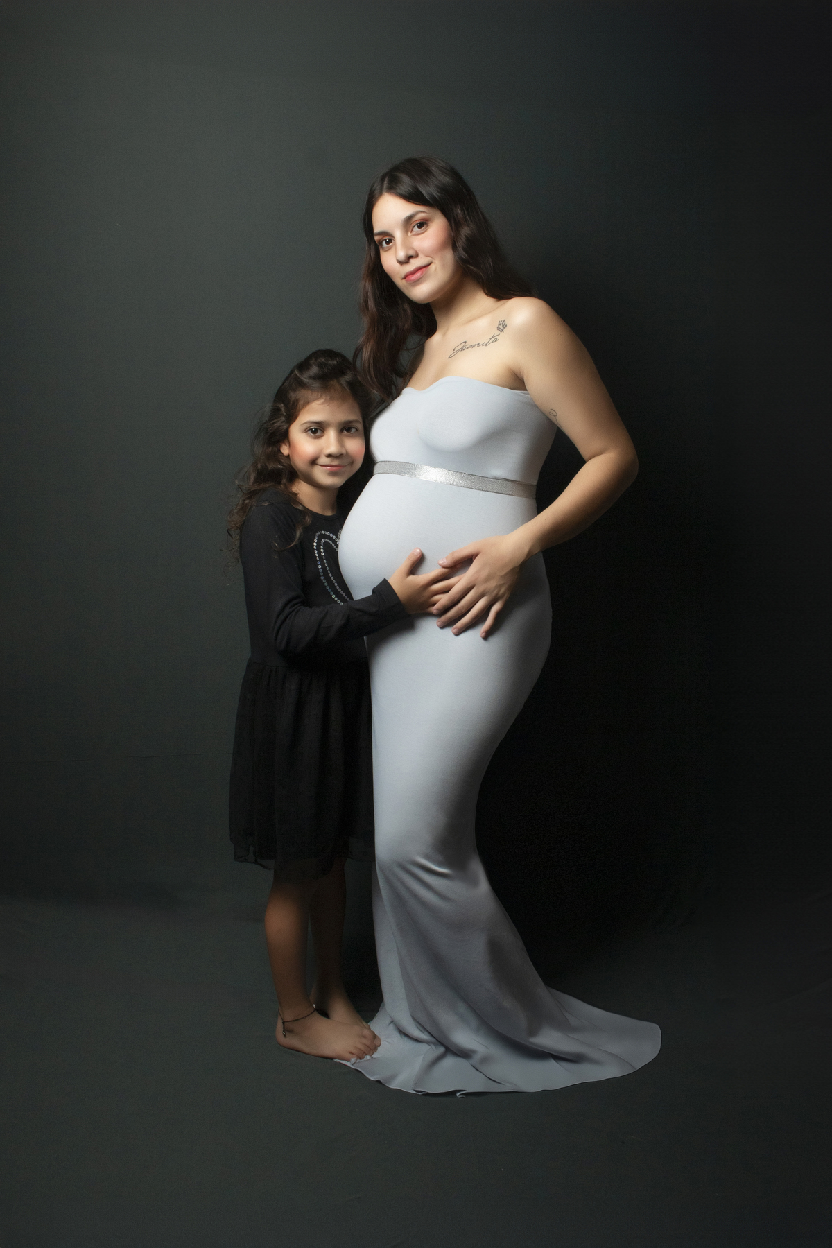 embarazada con vestido ajustado al cuerpo color celeste con una niña abrazando su panza , fondo liso tono gris azulado