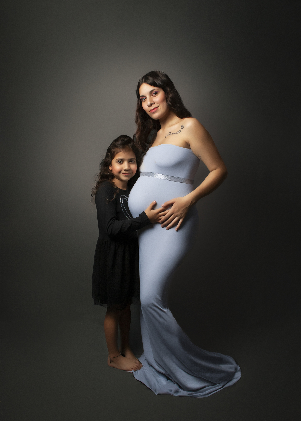 embarazada con vestido ajustado al cuerpo color celeste con una niña abrazando su panza , fondo liso tono gris