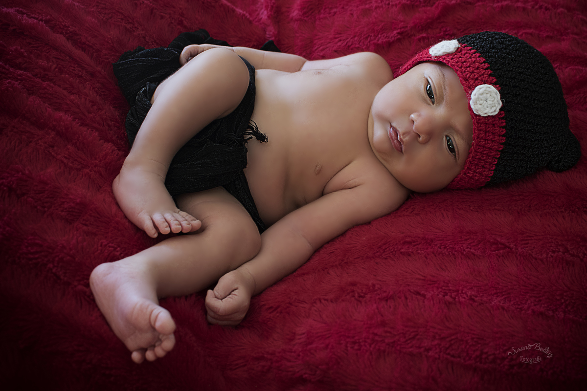 bebe completo con ojitos abiertos,  con gorrito negro, rojo y blanco envuelto con paño negro, sobre piel sintetica roja