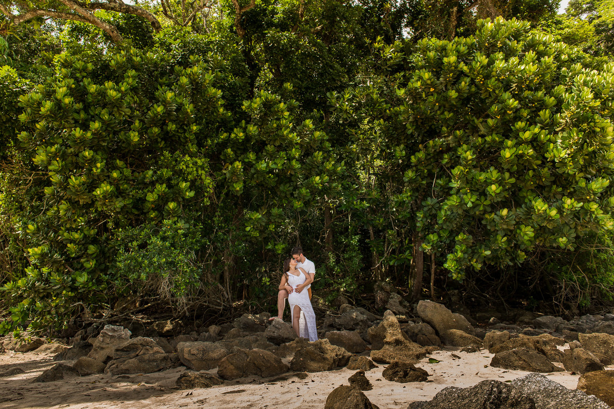 casal abraçados com arvore ao fundo na beira da praia