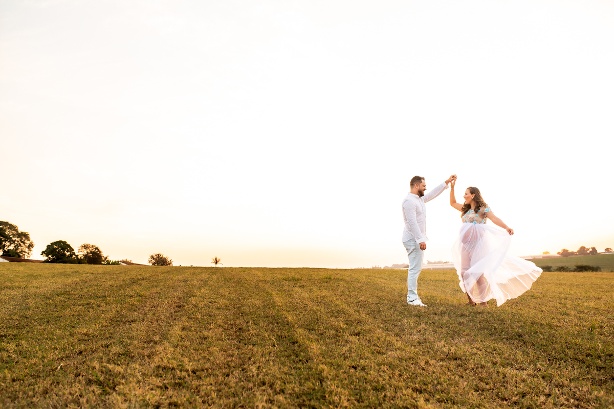 ensaio de gestante com casal dançando e sorrindo em campo de feno com o por do sol ao fundo em holambra cidade das flores