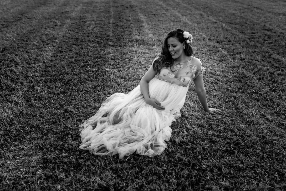 ensaio de gestante com com mulher sentada no chão de vestido sorrindo em campo de feno com o por do sol ao fundo em holambra cidade das flores