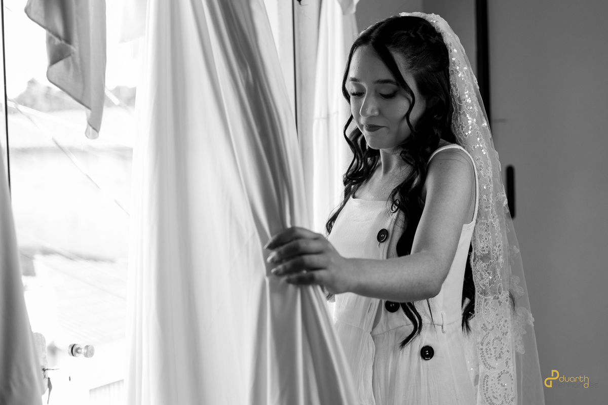 Noiva Mariana olhando para cortina com véu, momento contemplativo e de espera antes do casamento, foto P&B.