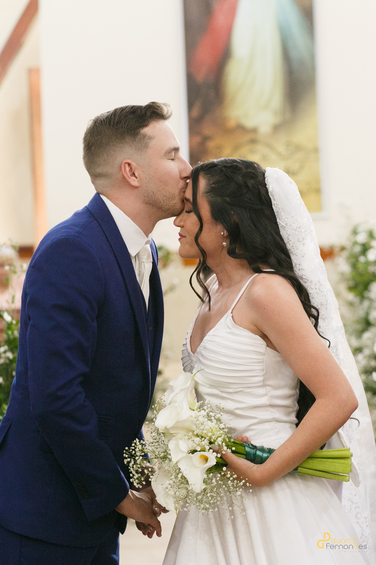 oivo Lucas beijando testa da noiva Mariana na igreja, buquê de noiva branco, casamento emocionante em Turmalina, MG.