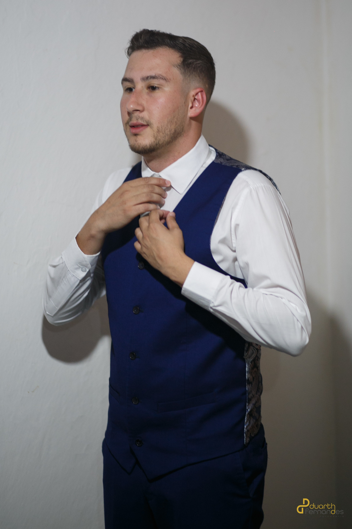 Noivo Lucas ajustando sua gravata branca com colete azul marinho e camisa branca, em pose elegante durante a preparação para o casamento