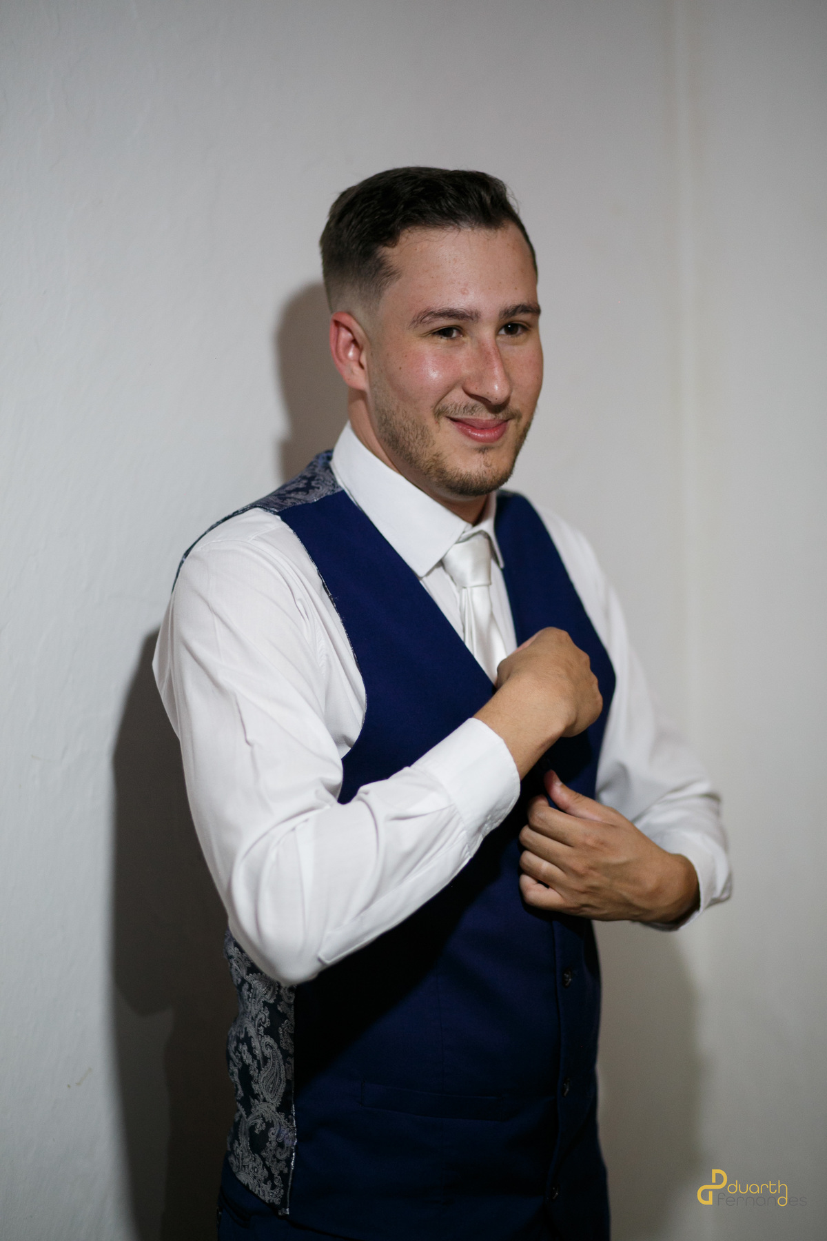 Noivo Lucas sorrindo enquanto ajusta sua gravata branca, vestindo colete azul marinho, em um momento descontraído e feliz de preparação para o casamento