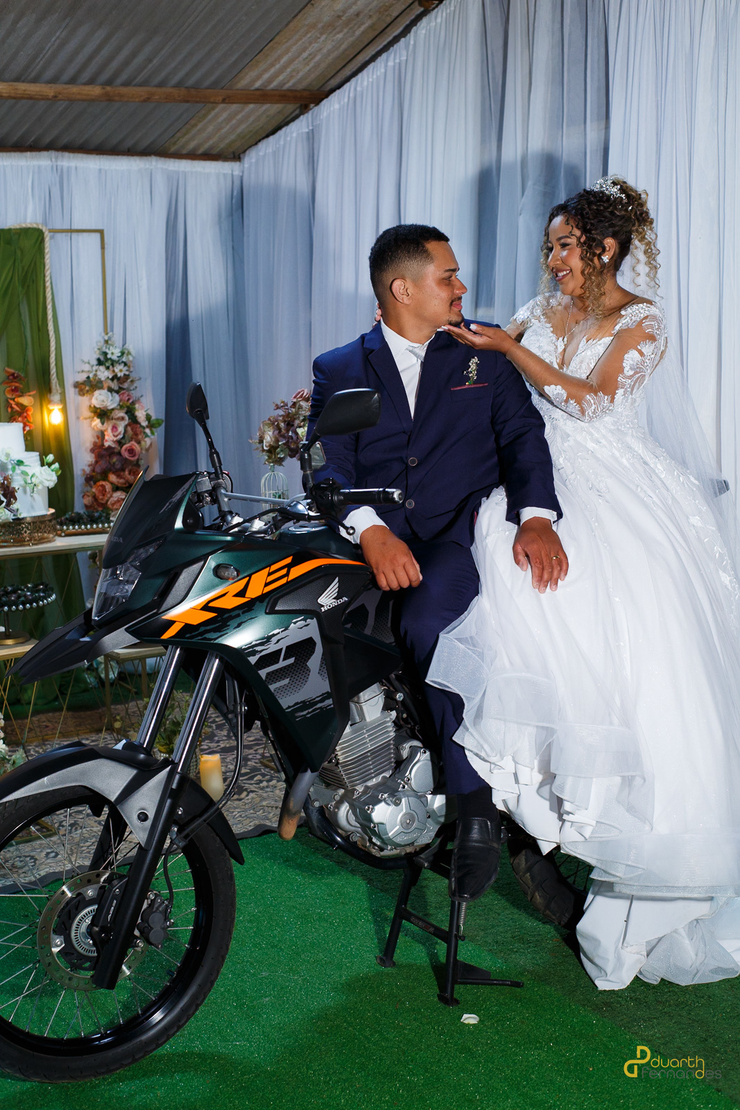 Edione e Emerson posando com uma moto XRE 300, em um cenário de decoração de casamento.
