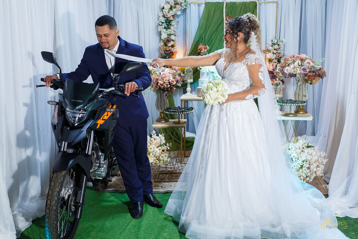 Edione e Emerson posando com uma moto XRE 300, em um cenário de decoração de casamento.