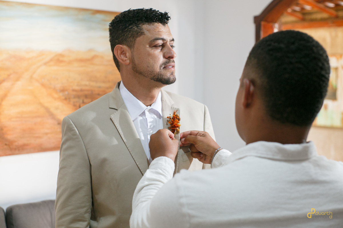Leandro, o noivo, sendo preparado por um amigo, recebendo o boutonnière. A imagem captura a emoção e os detalhes da preparação do casamento, com a arte no fundo complementando o momento.