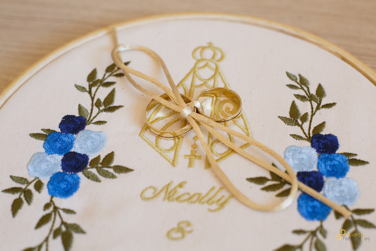 Alianças de ouro do casamento de Nicolly e Marcos descansando sobre um bastidor de bordado personalizado com uma imagem religiosa e flores azuis. Detalhe delicado e cheio de significado.