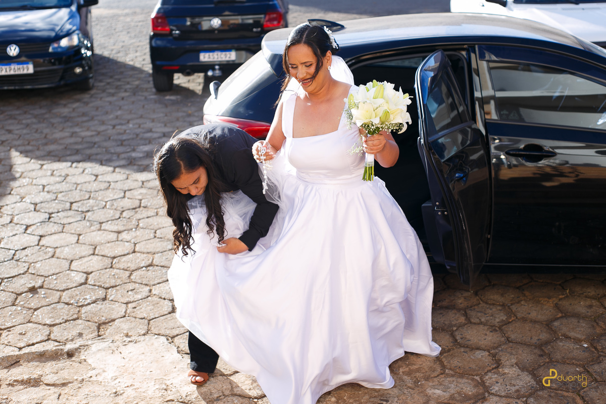 A noiva Nicolly chega ao casamento e recebe ajuda para arrumar o vestido ao descer do carro de noiva. Detalhes reais capturados por fotógrafo de casamento em Itamarandiba.