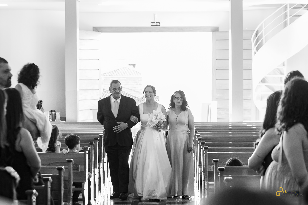 Entrada emocionante da noiva de braços dados com seus pais na igreja. Fotografia em preto e branco registrando a tradição e a emoção do casamento em Itamarandiba.