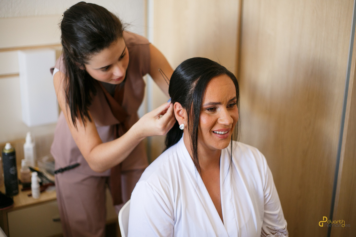 Noiva Nicolly sorrindo enquanto a cabeleireira finaliza o penteado com grampos. Um momento descontraído e feliz de preparação para o casamento em Itamarandiba.