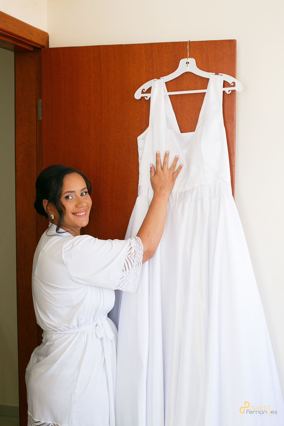 Noiva Nicolly radiante em seu hobby branco, tocando seu vestido de noiva pendurado na porta. A expectativa do grande dia em Itamarandiba, Minas Gerais.