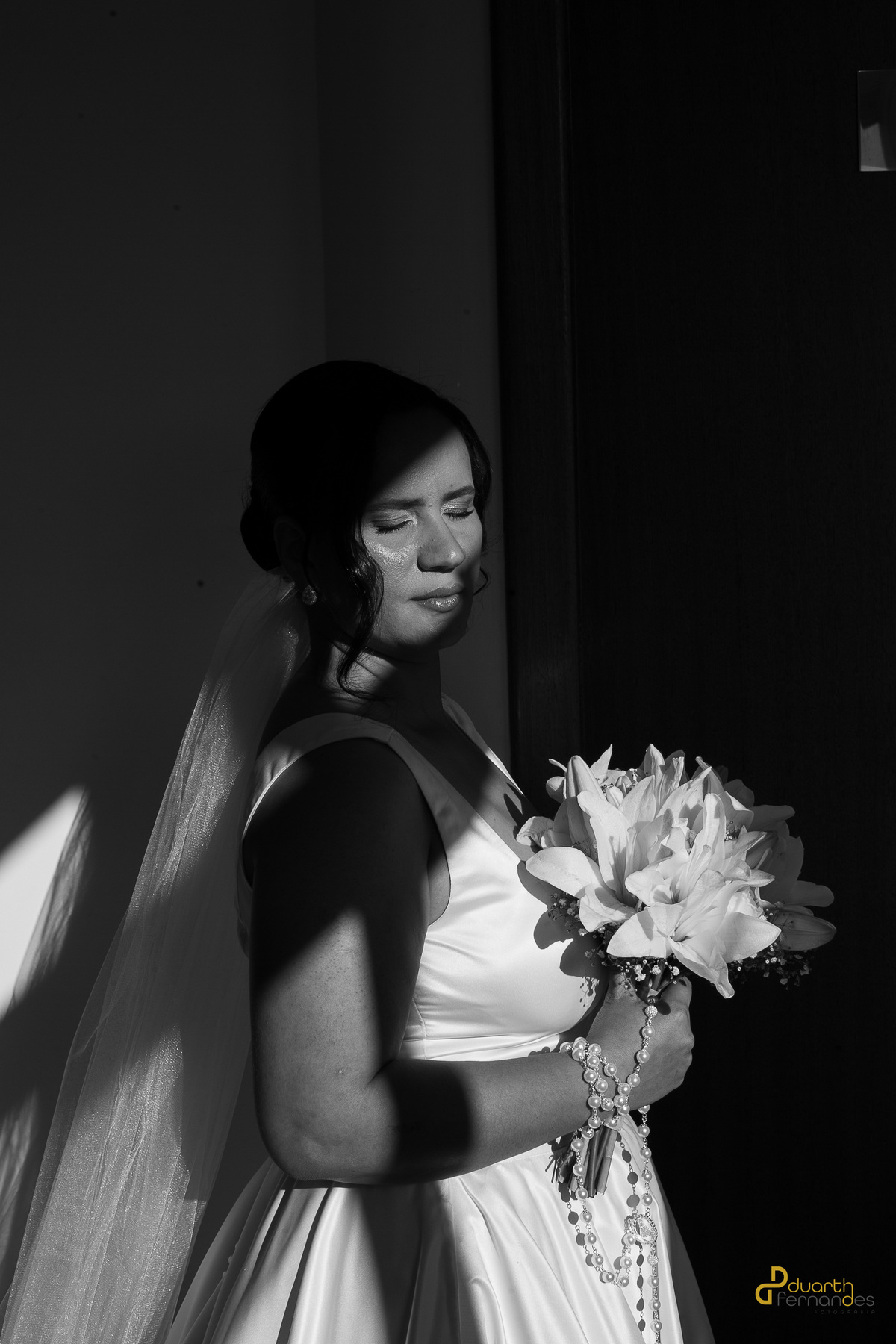 Noiva Nicolly em um momento íntimo, com os olhos fechados e a luz natural destacando sua beleza. Ela segura o buquê de lírios e o terço em preto e branco. Casamento em Itamarandiba.
