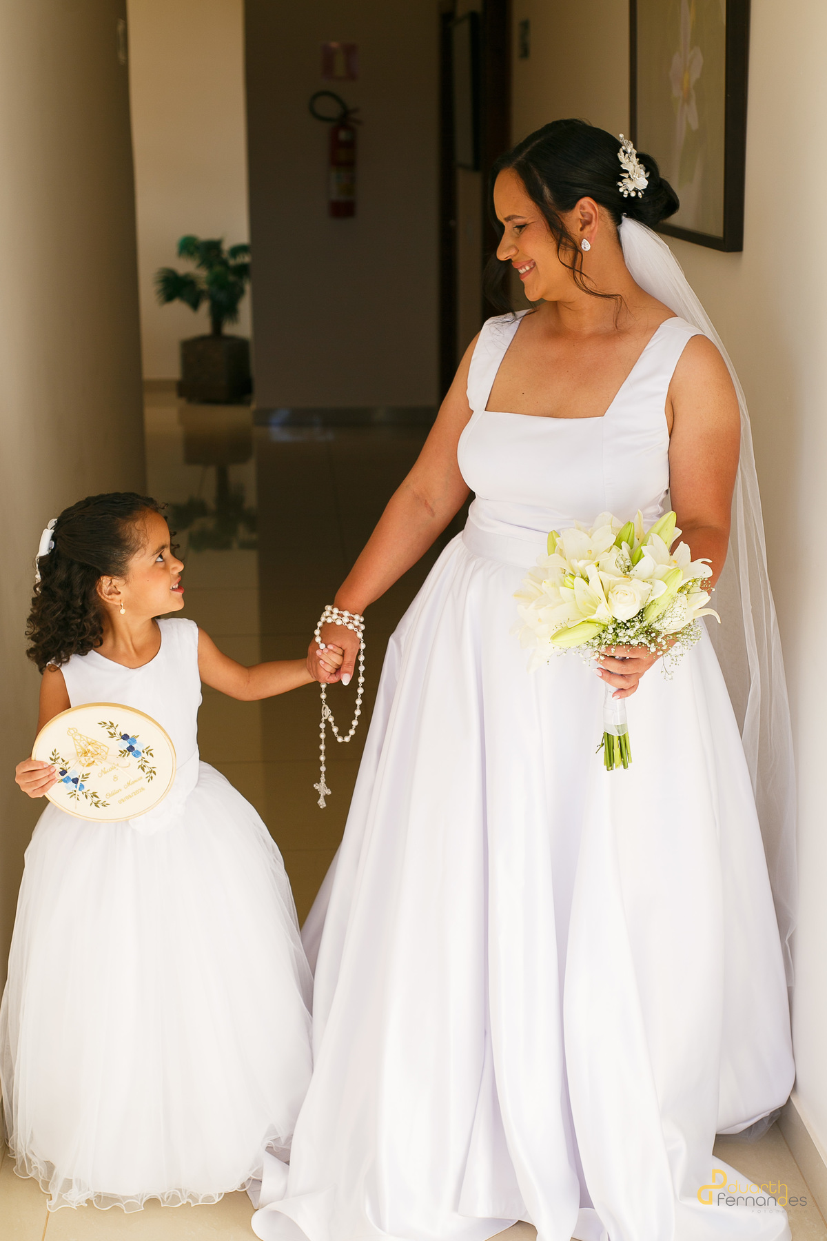 Noiva Nicolly de mãos dadas com a daminha de honra, que segura o bastidor de bordado personalizado. Um momento ternura e cumplicidade antes da cerimônia em Itamarandiba.