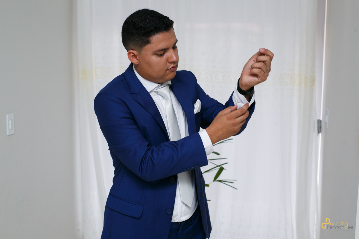 Noivo Arthur ajustando as abotoaduras da camisa branca sob o terno azul durante os preparativos do casamento. Foto realizada por fotógrafo em Aricanduva, MG.