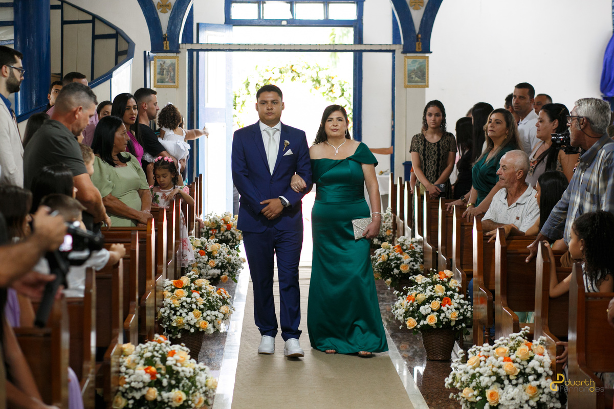 Entrada do noivo Arthur na igreja de braços dados com sua mãe, caminhando pelo tapete ladeado por arranjos de flores. Cerimônia de casamento em Aricanduva.