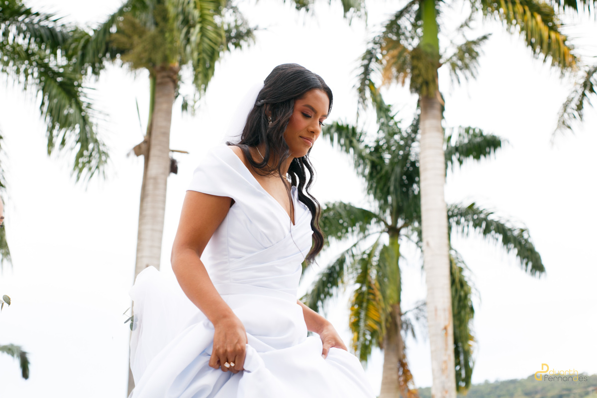 Noiva Gardênia com seu longo vestido branco e véu na entrada da igreja, ao lado do pai, emoldurada por um arco de flores coloridas no casamento em Aricanduva.