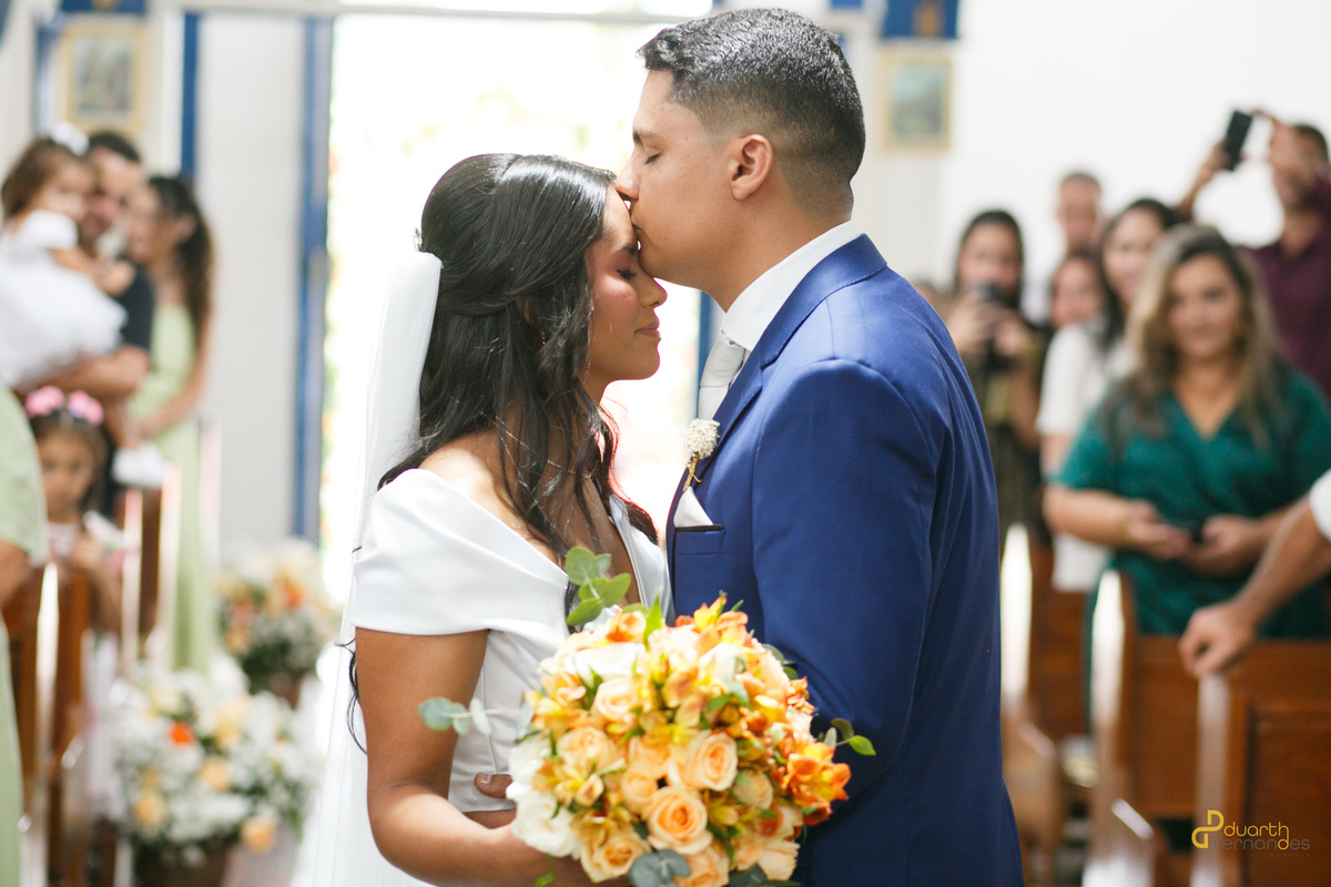 O noivo Arthur dá um beijo carinhoso na testa da noiva Gardênia no altar da igreja. Ela segura o buquê de rosas coloridas. Momento capturado por fotógrafo de casamento em Aricanduva.